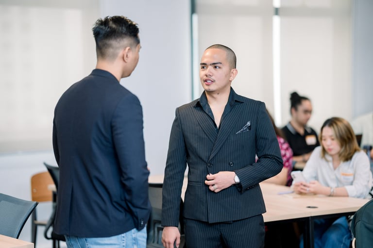 Two men having a conversation in a professional office setting with other colleagues seated in the background