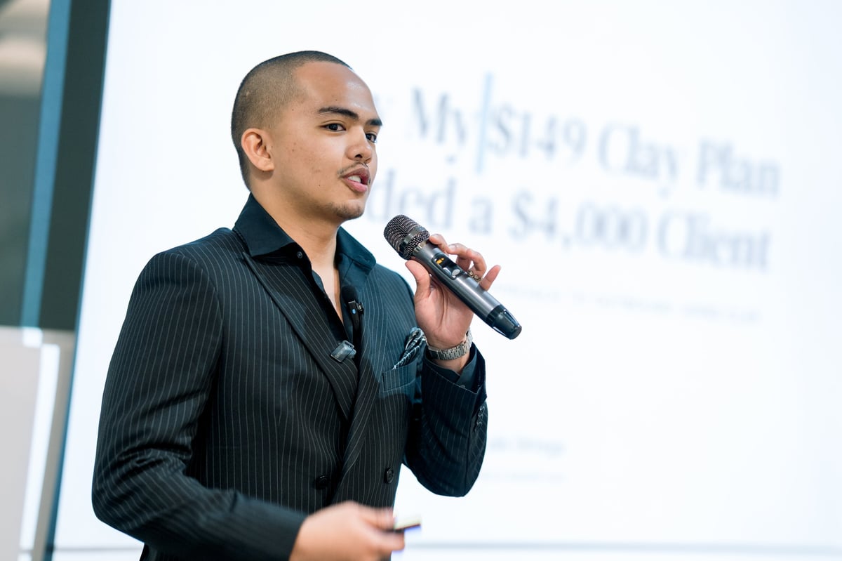 Man in dark pinstripe suit speaking into microphone at presentation with projection screen in background